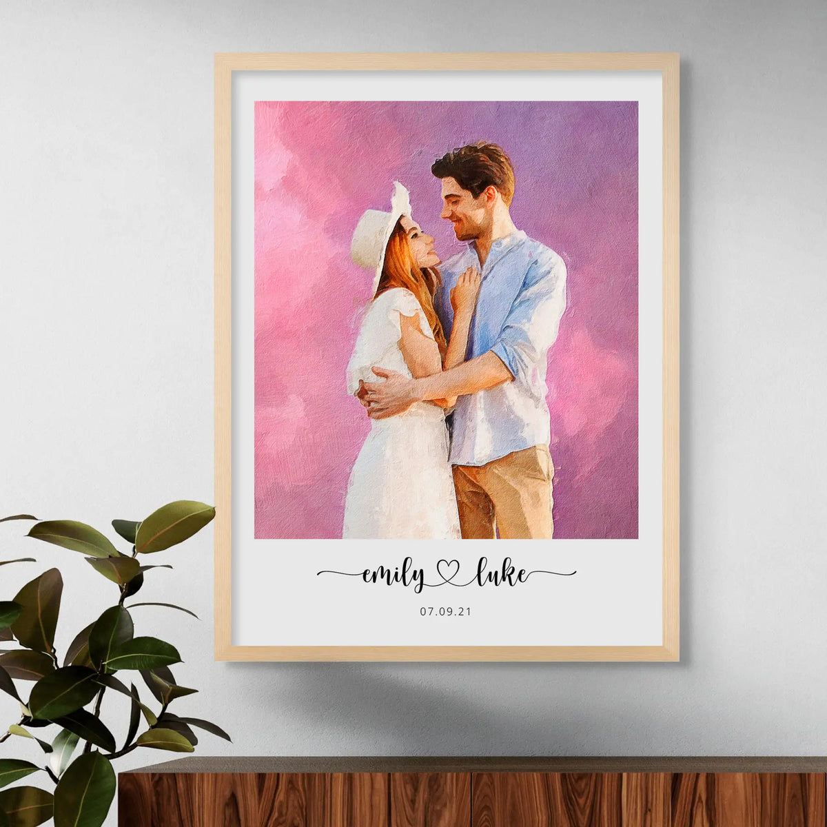 Oil-style portrait of a couple embracing on a pink and purple backdrop, in a light wood frame beside a leafy plant