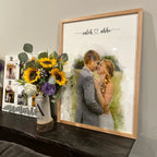 Customer photo of a watercolor-style portrait of a couple in a light oak frame, on a dark shelf beside sunflowers