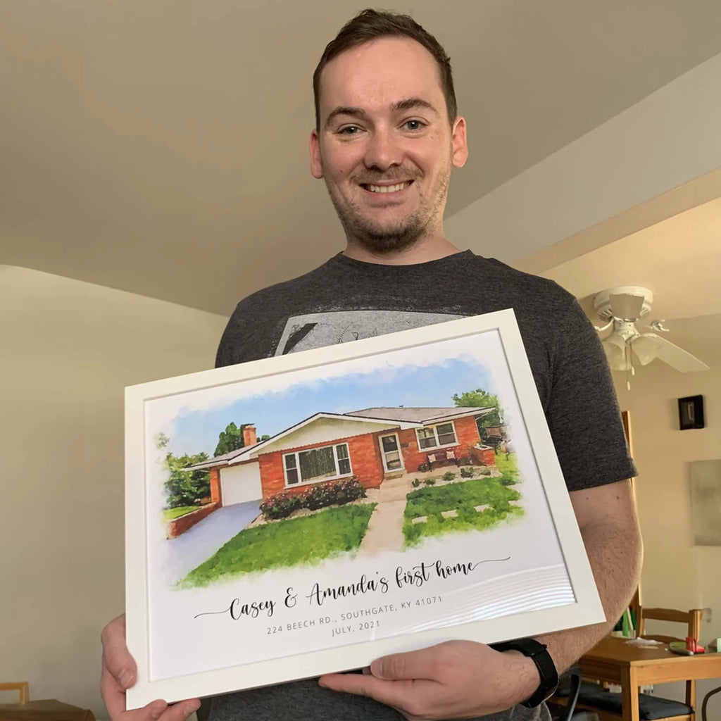A smiling man holds a white-framed watercolor-style house portrait of a brick bungalow, titled 'Casey & Amanda's first home'