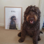 A curly-coated brown dog sits beside a framed watercolor-style pet portrait of a dark dog named 'Reilly'