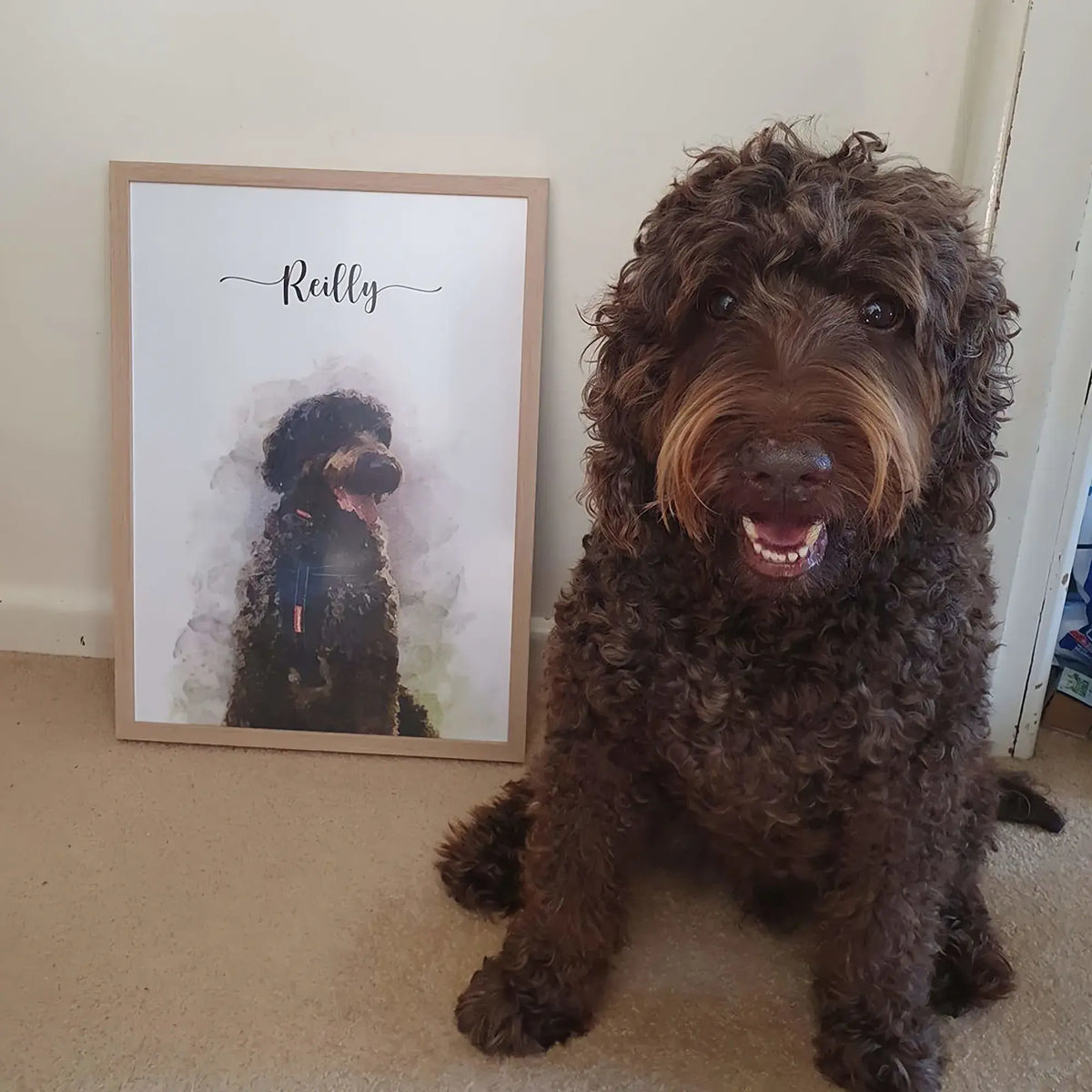 A curly-coated brown dog sits beside a framed watercolor-style pet portrait of a dark dog named 'Reilly'