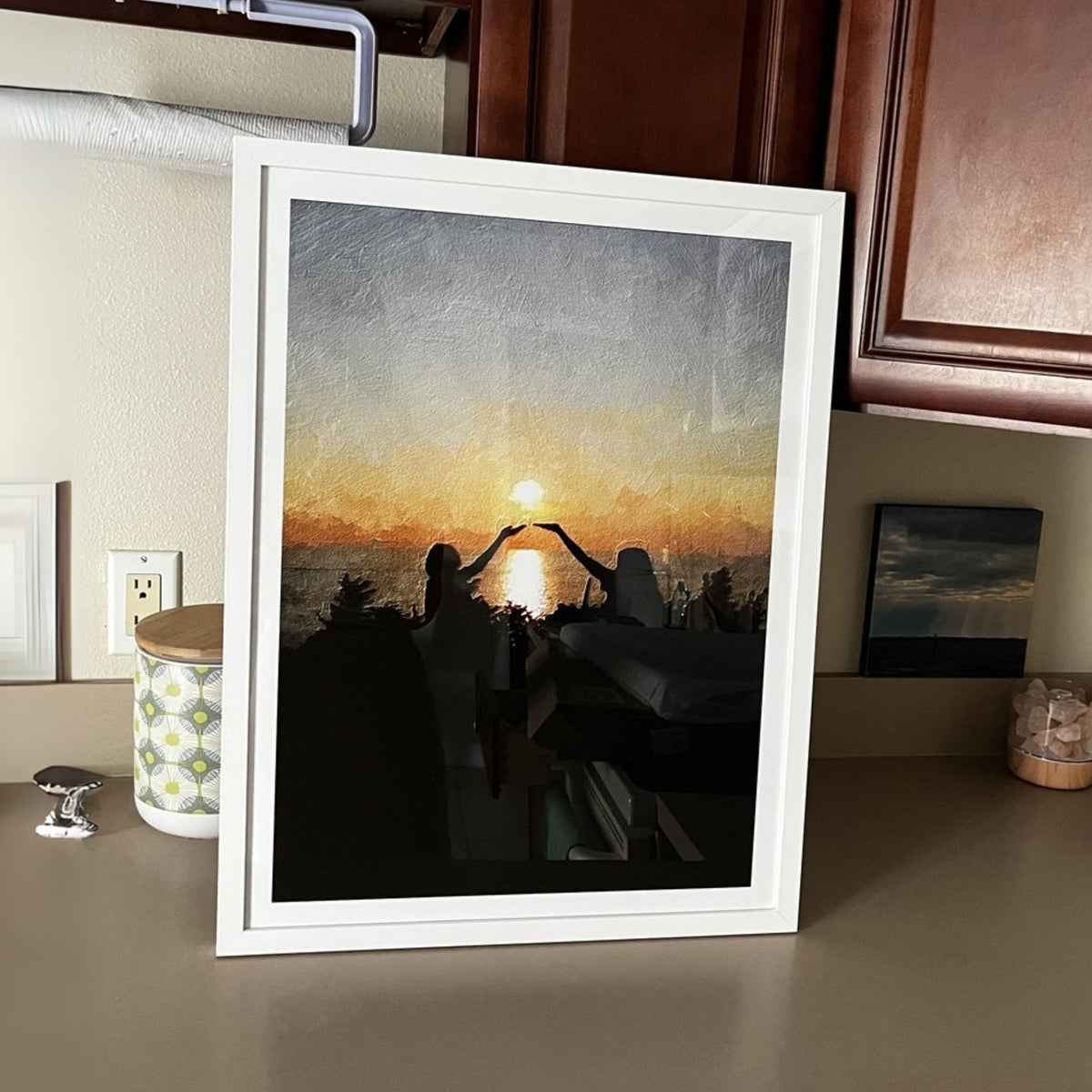 White-framed oil-style portrait of two silhouetted people raising their hands toward a setting sun, on a counter