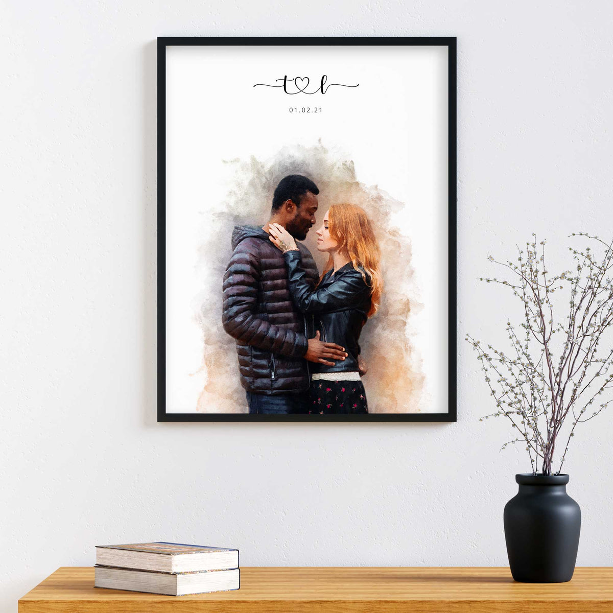 Watercolor-style portrait of a couple embracing, in a black frame above a wood table with books and a vase of twigs